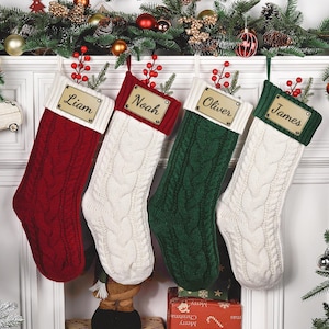 May include: Four cable-knit Christmas stockings hanging in a row. The first is red with a white cuff, the second is white with a red cuff, the third is green with a white cuff, and the fourth is white with a green cuff. Each has a gold nameplate.