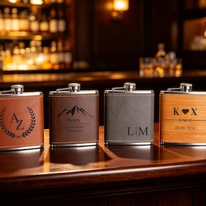 May include: Four rectangular metal flasks with leather-effect wraps in tan, brown, grey, and light wood-grain. Each features custom laser-engraved designs: initials with a laurel wreath, a mountain range with "Henry Groomsman 09-03-2026", initials "L|M", and "K ♥ X" with "08.09.2026".