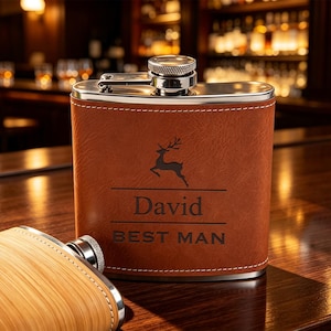 May include: A brown leather-wrapped stainless steel flask with a silver screw-top cap. The front features a black engraved graphic of a leaping deer above the text David and BEST MAN, separated by horizontal lines. A second flask is partially visible.