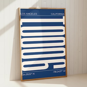 Puede incluir: Un cuadro rectangular con marco que presenta un fondo azul profundo con una tipografía grande, negrita y de color crema que dice LOS ANGELES CALIFORNIA. El texto incluye las coordenadas 34.0522 N y -118.2437 W en una fuente sans-serif.