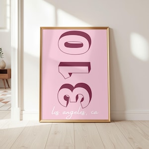 May include: A vertical rectangular wall art print in a gold frame featuring a light pink background with the large, bold, dark maroon 3D-style numbers 310 stacked vertically. Below the numbers, the text los angeles, ca is written in white.