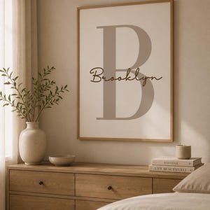 Pode incluir: Uma impressão de arte de parede emoldurada com uma letra B grande em cinza claro sobre um fundo creme. O nome Brooklyn está escrito em uma caligrafia cursiva marrom escura no meio da letra. A moldura é feita de madeira clara.