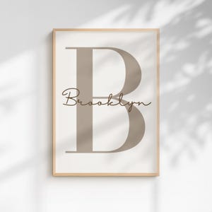 May include: A rectangular wall art print in a light wood frame featuring a large, centered, taupe-colored serif letter B. Overlaid across the center of the letter is the name Brooklyn written in a dark brown, flowing, cursive script font.
