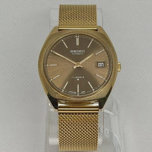 May include: A gold-tone Seiko automatic wristwatch featuring a round brown dial with gold-tone hour markers and hands. The watch includes a date window at the three o'clock position and a gold-tone mesh metal strap with a secure clasp mechanism.