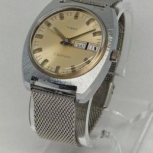 Könnte beinhalten: Eine Timex Electronic Armbanduhr mit rundem, goldfarbenem Zifferblatt, Strichindizes, silberfarbenen Zeigern und einem Datumsfenster, das SAT und die Zahl 11 anzeigt. Die Uhr hat ein gebürstetes, silberfarbenes Metallgehäuse und ein Edelstahl-Mesh-Armband.