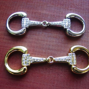 Puede incluir: Dos abalorios de metal en forma de bocado de caballo con detalles de strass transparentes. Un abalorio es plateado y el otro es dorado.