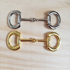 May include: Two metal clasps, one silver and one gold, with a D-ring shape and a connecting bar. The clasps are on a wooden surface.