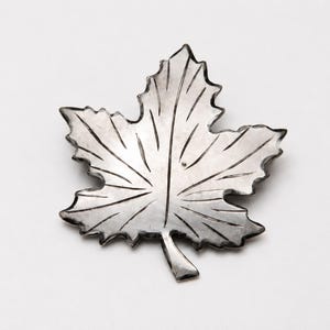 May include: A silver-toned metal brooch shaped like a maple leaf. The surface features etched lines radiating from the central stem to the edges of the leaf, creating a detailed vein pattern. The edges are serrated and the finish is metallic.