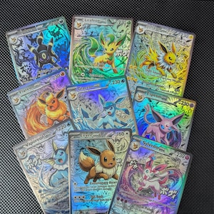 May include: A collection of eight holographic Pokemon trading cards featuring Eevee and its various evolutions including Umbreon, Leafeon, Jolteon, Flareon, Glaceon, Espeon, Vaporeon, and Sylveon, each displaying colorful character illustrations and text.