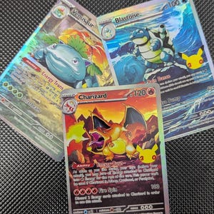 May include: Three holographic trading cards featuring Venusaur, Blastoise, and Charizard. Each card displays the character name, HP values, ability text, and attack details. The Charizard card is centered, with the Venusaur and Blastoise cards positioned behind it.