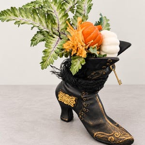 May include: A black boot-shaped planter with gold scrollwork accents, wrapped in black netting. It contains a green fern, an orange pumpkin, and a white pumpkin. A small gold key hangs from the top eyelet of the boot, which features a high heel.
