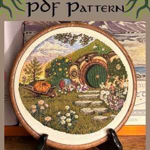 May include: A circular embroidery hoop containing a detailed fabric stitch of a round green door set into a grassy hill. The scene includes a stone path, a wheelbarrow with a pumpkin, colorful flowers, and a blue sky with clouds. Text reads PDF PATTERN.