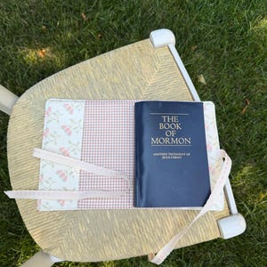 May include: A dark blue book titled The Book of Mormon Another Testament of Jesus Christ resting on a fabric cover. The cover features a light pink and white gingham pattern, a cream panel with a small floral print, and two thin lace ties.