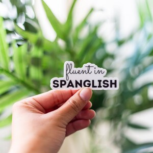 May include: A white sticker with black text that reads "fluent in Spanglish". The sticker is being held by a hand against a blurred background of green plants.