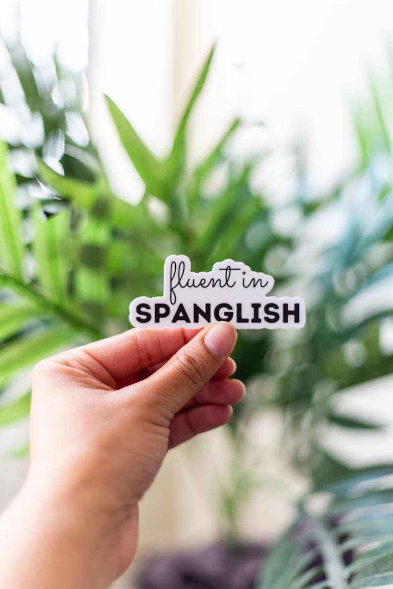 May include: A white sticker with black text that reads "fluent in Spanglish". The sticker is being held by a hand against a blurred background of green plants.