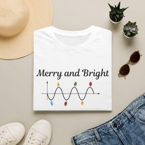 Puede incluir: Una camiseta blanca con el texto negro Merry and Bright sobre un gráfico de onda sinusoidal negro. El gráfico incluye pequeñas bombillas navideñas de colores rojo, amarillo, azul y verde situadas en los picos y valles de la onda.