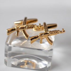 May include: A pair of gold-toned cufflinks shaped like AK-47 assault rifles. The cufflinks are displayed on a clear acrylic stand.