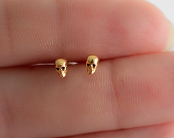 Puede incluir: Un par de pendientes de botón de calavera de oro. Los pendientes son pequeños y delicados, y son perfectos para agregar un toque de estilo atrevido a cualquier atuendo.