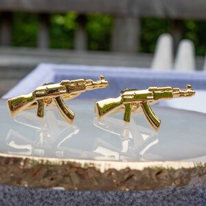 May include: Two gold-coloured miniature AK-47 assault rifle cufflinks. The cufflinks are on a white surface.