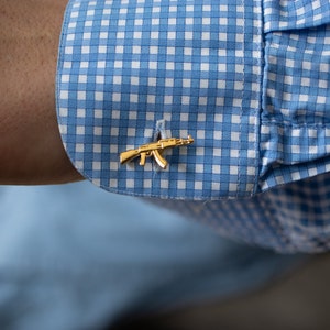 May include: Gold cufflinks in the shape of a rifle, on a blue and white checked shirt cuff.