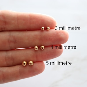 Puede incluir: Tres pares de pendientes de bola de oro en diferentes tamaños: 3 mm, 4 mm y 5 mm. Los pendientes se muestran en una mano.