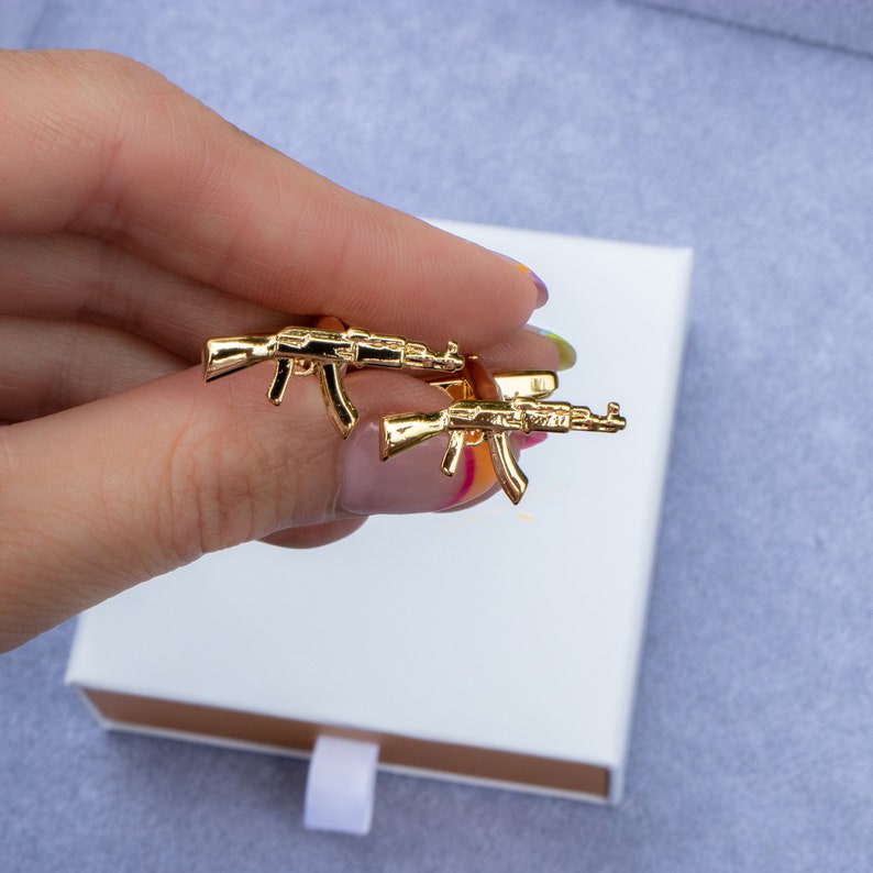 May include: A pair of gold-tone cufflinks shaped like AK-47 assault rifles. The cufflinks are displayed on a white box.