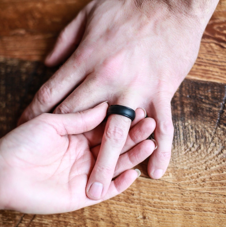 Op de afbeelding: Een close-up van twee handen, waarvan er &eacute;&eacute;n een zwarte siliconen ring draagt. De ring is simpel en modern, met een gladde, matte afwerking.