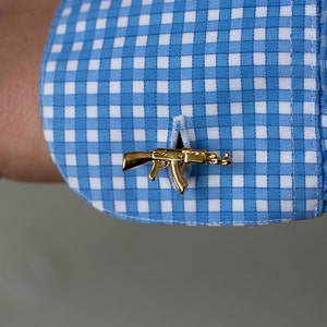 May include: Gold cufflinks in the shape of a rifle, on a blue and white gingham shirt.