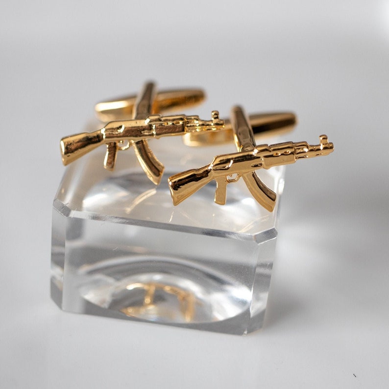 May include: A pair of gold-toned cufflinks shaped like AK-47 assault rifles. The cufflinks are displayed on a clear acrylic stand.