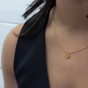 May include: A delicate gold necklace with a small, round pendant. The pendant has the letter "S" engraved on it. The necklace is worn against a black top, creating a contrast that highlights the jewelry. The chain is a fine, delicate design.