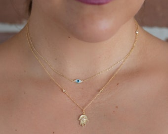 Könnte beinhalten: Eine goldene Kettenkette mit einem Hamsa-Hand-Anhänger und einem blauen bösen Auge. Die Kette ist mit zwei Ketten geschichtet, eine mit kleinen goldenen Perlen und die andere mit einer einfachen Kette.