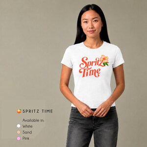 May include: A white short-sleeved t-shirt featuring the red text "Spritz Time" in a cursive font, accompanied by a small orange hibiscus flower graphic. The shirt is paired with dark gray jeans. Text below indicates color options: White, Sand, Pink.