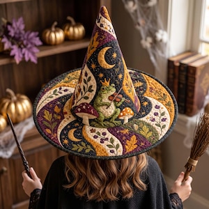 May include: A quilted witch hat featuring a central green frog wearing a crown and playing a violin, surrounded by mushrooms, crescent moons, stars, and botanical patterns in shades of purple, gold, black, and cream with a pointed crown.