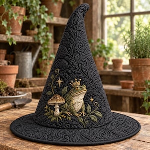 May include: A black pointed hat featuring an intricate quilted texture throughout. The front displays an embroidered design of a green frog wearing a gold crown, positioned next to a brown mushroom and surrounded by delicate leafy vines and stars.