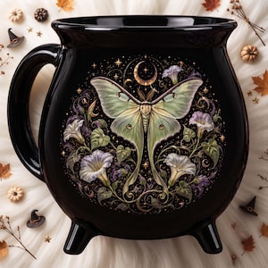 May include: A black ceramic cauldron-shaped mug featuring a central illustration of a pale green luna moth surrounded by white morning glory flowers, swirling vines, and a golden crescent moon with small stars against a dark background.