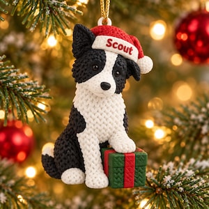 May include: A hanging ornament shaped like a black and white dog with a textured knit appearance. The dog wears a red Santa hat with the name Scout printed in red text. One paw rests on a green gift box with a red ribbon, also featuring a knit texture.