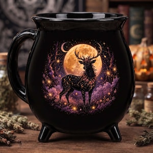 Puede incluir: Una taza negra con forma de caldero que presenta una ilustración circular de un ciervo con astas adornadas con flores. El ciervo se encuentra en un bosque morado bajo una gran luna naranja brillante con símbolos de media luna en su cuerpo.