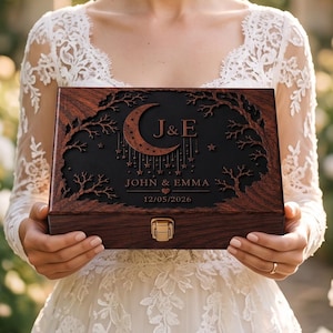 May include: A rectangular wooden keepsake box with a dark brown finish and a black lid featuring laser-cut details of a crescent moon, stars, and tree branches. The lid is engraved with the initials J & E, the names JOHN & EMMA, and the date 12/05/2026.