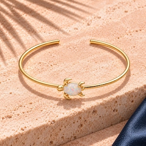 Peut inclure: Un bracelet jonc ouvert doré orné d'un motif central en forme de tortue. Le motif de la tortue est incrusté d'une pierre blanche lisse et irisée, semblable à de l'opale, révélant de subtils reflets colorés sur sa carapace ovale.