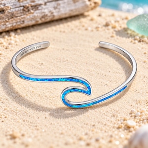 May include: A silver-toned cuff bracelet featuring a wave-shaped design inlaid with vibrant blue crushed opal. The metal surface is engraved with the text Always with you along the inner curve of the band, creating a sleek and polished appearance.