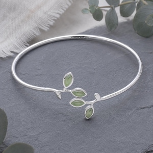 May include: A silver bangle bracelet featuring a branch motif with four light green, leaf-shaped stones. The metal band is engraved with the text Always with you. The design is open-ended, showcasing a polished metallic finish and smooth stones.