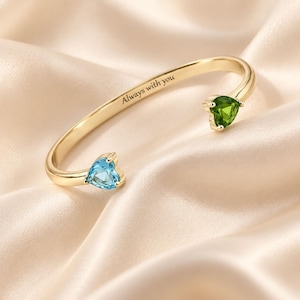 May include: A gold-toned open cuff bracelet featuring two heart-shaped gemstones, one light blue and one green, set at each end. The inner band is engraved with the text Always with you in a dark, clear font, creating a polished appearance.