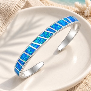 Puede incluir: Una pulsera tipo brazalete de tono plateado que presenta una serie de incrustaciones rectangulares de ópalo azul iridiscente. La pulsera tiene un acabado metálico pulido y un diseño abierto, mostrando un patrón vibrante y moteado en los segmentos de piedra azul.
