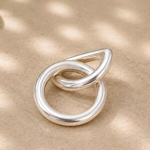 May include: A polished silver ring featuring two interlocking shapes, a circular band and a teardrop-shaped loop. The metal has a smooth, reflective surface with a rounded profile, creating a continuous, fluid design that connects the two distinct geometric forms.