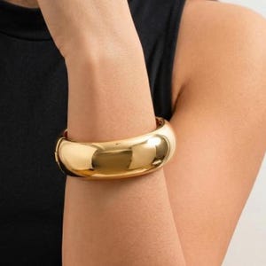 Puede incluir: Una pulsera tipo brazalete ancha de tono dorado pulido con una superficie lisa y redondeada y un acabado de alto brillo. La pulsera presenta un diseño de banda gruesa y sólida que rodea la muñeca, mostrando una estética limpia.