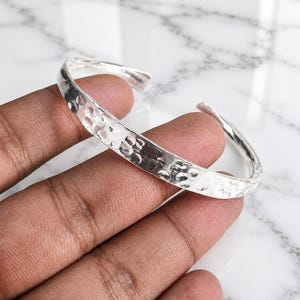 May include: A silver-colored cuff bracelet featuring a hammered, textured surface with multiple circular indentations. The metal band has an open design, allowing it to be slipped onto the wrist. The finish is reflective and polished.