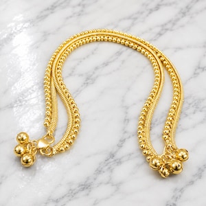 Op de afbeelding: Een goudkleurig enkelbandje met een dubbelstrengs ontwerp, bestaande uit een ketting met kleine bolvormige kralen en een platte, gevlochten schakelketting. Beide uiteinden zijn versierd met trosjes kleine, ronde, gepolijste goudkleurige belletjes.