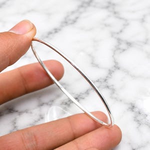 Op de afbeelding: Een dunne, gepolijste zilveren armband met een glad, afgerond oppervlak. De metalen band is cirkelvormig en heeft een reflecterende, metallic afwerking die het licht opvangt, wat zorgt voor een eenvoudige en strakke uitstraling van dit sieraad.