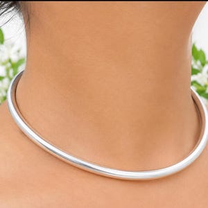May include: A smooth, polished silver-tone metal choker necklace with a rounded, tubular design. The necklace sits firmly around the neck, featuring a continuous, seamless band with a reflective, high-shine finish that catches the light.