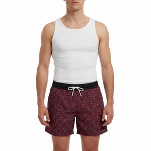 May include: A white ribbed tank top paired with dark maroon swim trunks featuring a repeating floral pattern in shades of pink and green. The shorts have a solid black elastic waistband and a white drawstring cord tied at the center front.